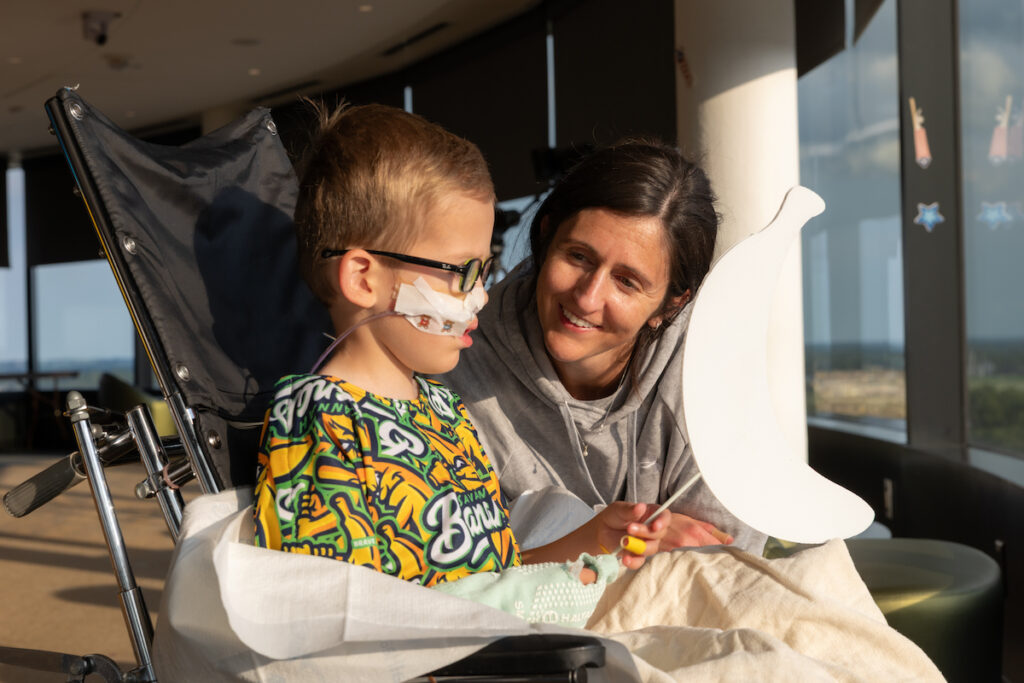 A young child with medical equipment sits in a wheelchair beside an adult who smiles while holding a large white banana-shaped object.