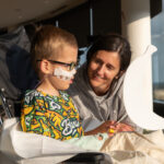A young child with medical equipment sits in a wheelchair beside an adult who smiles while holding a large white banana-shaped object.