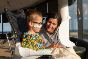 A young child with medical equipment sits in a wheelchair beside an adult who smiles while holding a large white banana-shaped object.